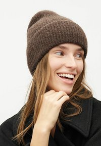 Căciulă din tricot cu nervuri, de culoare maro, realizată dintr-un material moale și texturat. Are un manșon pliat și un vârf ușor căzut, oferind o croială confortabilă.