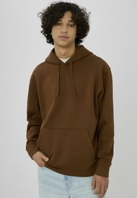 Jeune homme aux cheveux bouclés portant un sweat à capuche marron et un jean bleu clair, debout avec une main dans la poche, sur un fond uni.