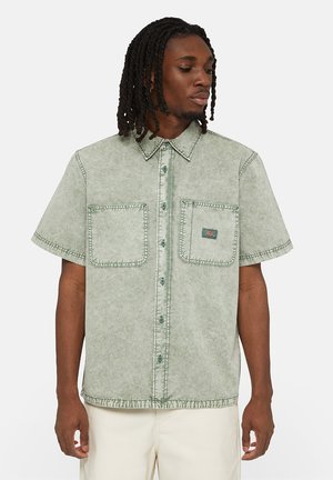 Camisa de manga corta en denim verde claro con dos bolsillos en el pecho, costuras visibles y un cierre de botones en la parte delantera. La textura parece descolorida.