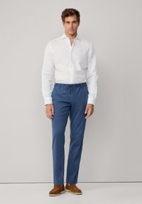 Man in een wit langarmshirt, blauwe broek met trekkoord en bruin suède schoenen die tegen een eenvoudige lichte achtergrond staat.