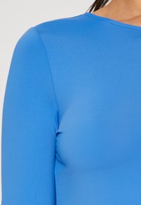 Nærbillede af en person iført en langærmet, lyseblå tætsiddende top med rund hals ud mod en ensfarvet baggrund.