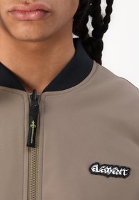 Khaki bomber jacket with black ribbed collar and zipper. Features a contrasting logo patch and decorative zipper pull with green detailing.
