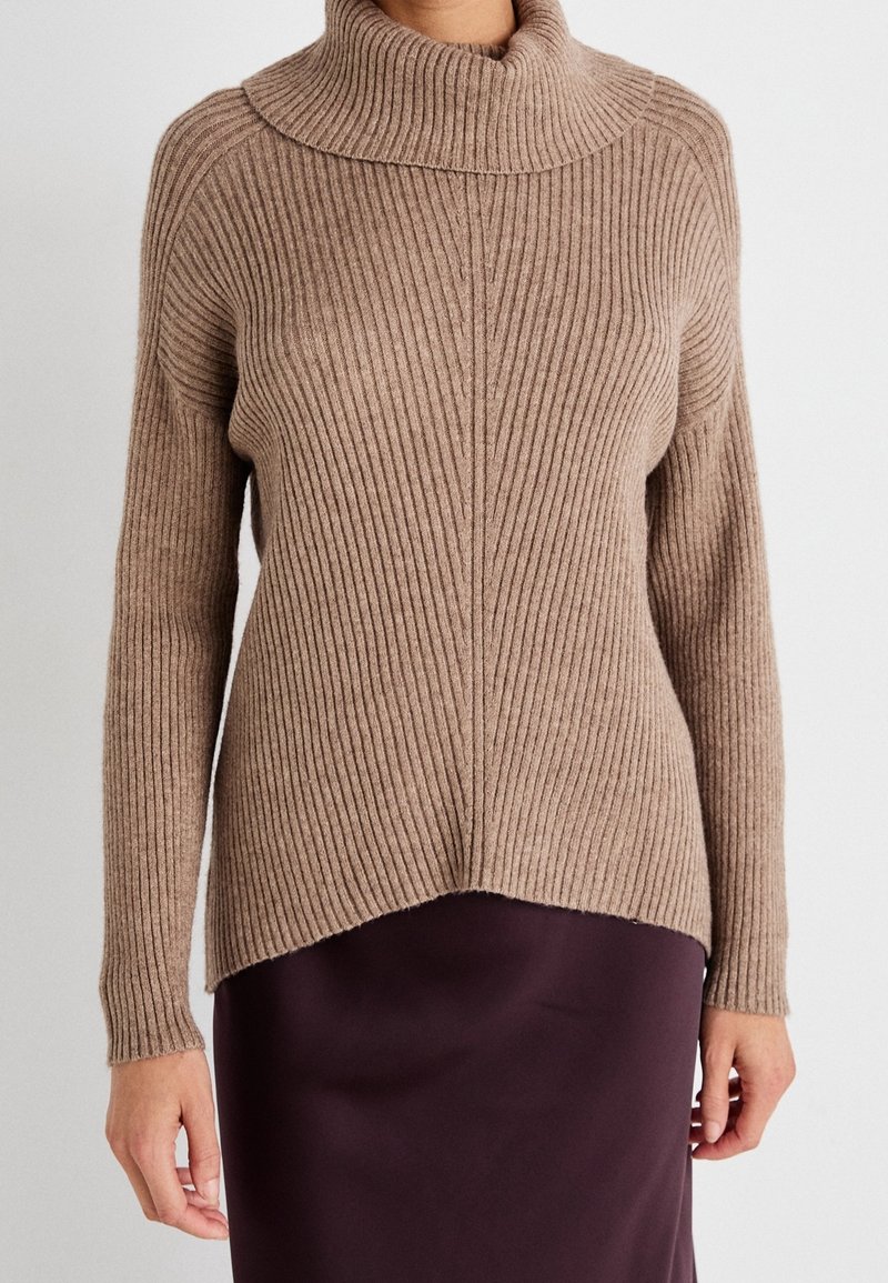 Pull en tricot côtelé marron avec un col haut et des manches longues. L'ourlet est légèrement asymétrique, associé à une jupe violette foncée.