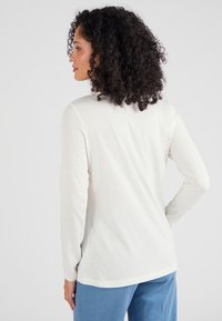 Lichtbeige lange mouw top gemaakt van zacht, lichtgewicht stof. Heeft een ronde halslijn en subtiele naden aan de achterkant. Gecombineerd met blauwe broek.