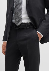 Dark suit with a textured herringbone tie. The belt is black leather. Fabric is smooth with a slight sheen; tailored fit visible.