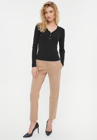 Czarna, długosleeve ribbed bluzka z dekoltem w serek i guzikami, w połączeniu z beżowymi spodniami w kant i czarnymi szpilkami, na jednolitym tle.