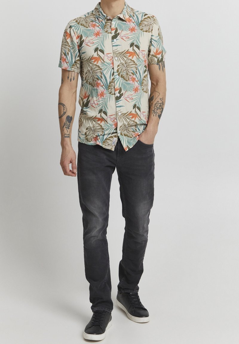 Homme portant une chemise à manches courtes imprimée de fleurs, un jean noir, des baskets noires, debout avec une main dans une poche devant un fond uni.