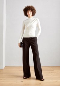 Cremefarvet langærmet top med draperet stofdetalje, parret med højtaljede sorte bredsmyndede bukser. Model holder en lille sort clutch.