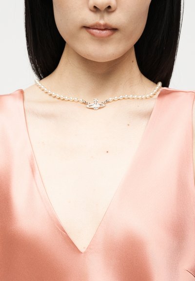 Γυναίκα που φοράει ένα κολιέ από μαργαριτάρια με ασημένιο κρεμαστό σφαιρίδιο, ντυμένη με ένα ροζ σατέν τοπ με V-λαιμόκοψη, εμφανίζοντας τον λαιμό και την περιοχή της κλείδας.