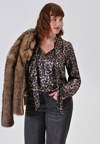 Donna che indossa una blusa con stampa leopardata e jeans neri, tiene un cappotto in pelliccia sintetica marrone sopra la spalla, guarda verso destra.