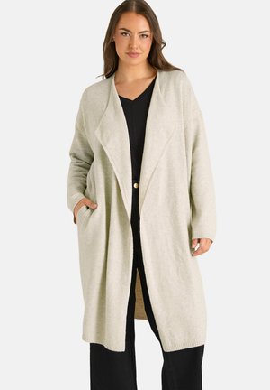 Woman wearing a light beige open-front long cardigan with pockets over a black V-neck top and black pants, standing against a plain background.