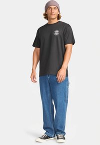 Schwarzes Baumwoll-T-Shirt mit rundem Logo-Druck auf der Brust, kombiniert mit hellblauen baggy Denim-Jeans und schwarzen Canvas-Sneakers.