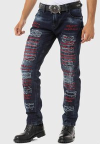 Blaue Jeans aus Denim mit Rissen und fransigen Kanten, versehen mit roten Textakzenten. Gerades Bein-Design kombiniert mit schwarzen Stiefeln und einem Gürtel.