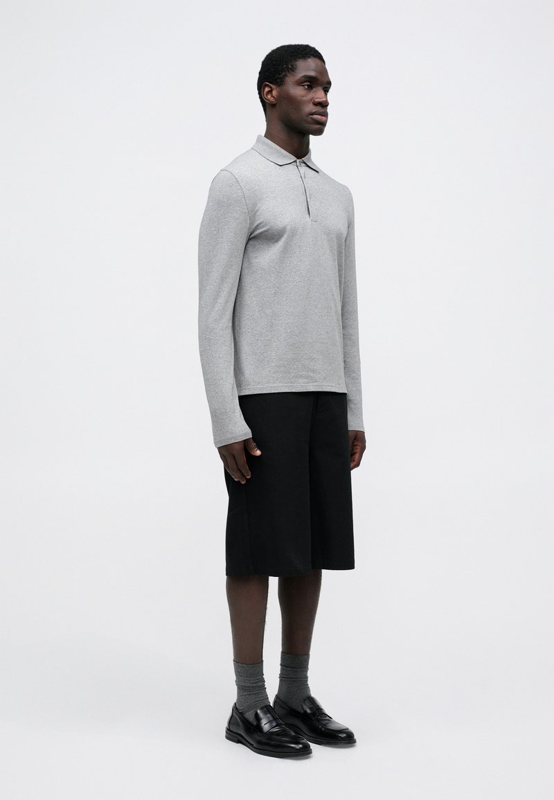 Polo grigio a maniche lunghe con colletto, abbinato a pantaloni corti neri fino al ginocchio. Completato con calzini grigi e mocassini neri.