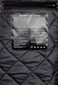 Czarna etykieta z instrukcjami dotyczącymi pielęgnacji pikowanej tkaniny, z wyraźnym białym tekstem i ikonami dotyczącymi prania, suszenia i zaleceń dotyczących prasowania.