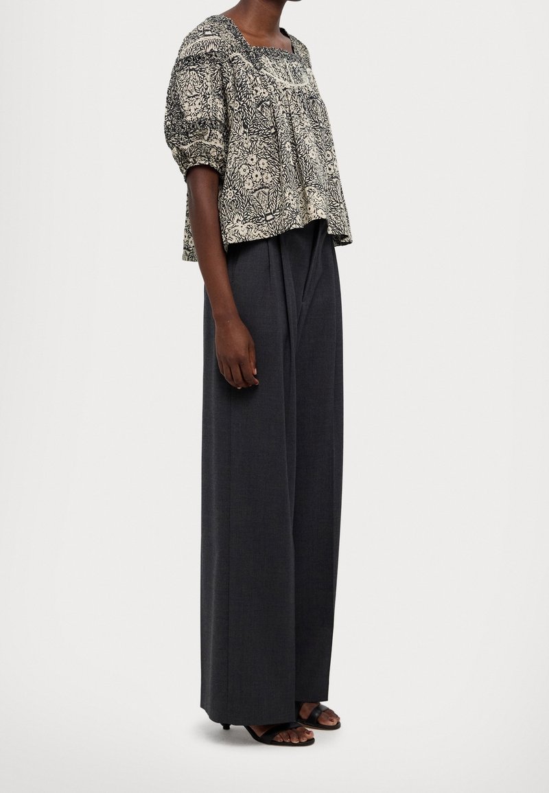 Femme portant un chemisier ample à motifs avec des manches bouffantes et un pantalon large gris foncé avec des sandales noires à bout ouvert.