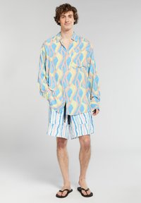 Camicia button-up leggera e colorata con un motivo ondulato in rosa e blu; abbinata a pantaloni corti a righe e ciabatte nere.