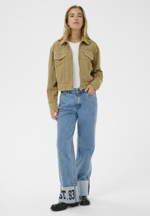 Kvinde iført nittebesat tan-jakke over hvid skjorte, vidde ben blå jeans med opviklede manchetter og sorte støvler, stående mod en hvid baggrund.