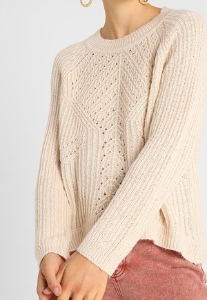 Pull en maille beige clair avec un motif en diamant texturé, des poignets côtelés et un col rond. Présente un détail de point ouvert.
