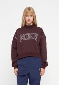 Bordo kratki dukserica s visokim ovratnikom, s bijelim logotipom "NIKE" i kontrastnim šavovima. Uparen s tamnoplavljim hlačama.