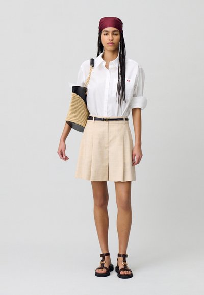 Camicia bianca con bottoni, pantaloni corti beige a pieghe, cintura marrone, sandali marroni con cinturini e borsa in paglia intrecciata con dettagli neri.