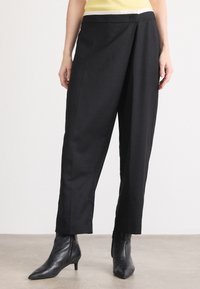 Pantalons noirs ajustés avec une coupe loose, présentant un devant plat, des poches latérales et une texture lisse. Associés à des bottines noires à talons pointus.