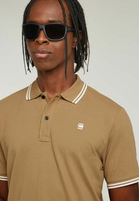Polo shirt marrone con colletto a righe bianche, placket a due bottoni e piccolo logo. Il modello indossa occhiali da sole neri; lo sfondo è neutro.