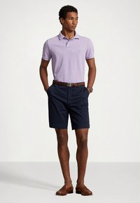 Πόλο μπλουζάκι λιλά με κοντά μανίκια και πράσινο λογότυπο, συνδυασμένο με σορτς σε ναυτικό μπλε και καφέ δερμάτινα παπούτσια. Η καφέ ζώνη ολοκληρώνει το σύνολο.