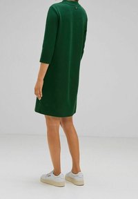 Grön klänning med trekvartslånga ärmar, lätt A-linjeformad, tygtextur synlig. Knappdetalj på baksidan. Matchad med vita sneakers.