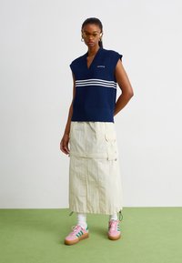 Μπλέ αμάνικο πλεκτό τοπ με λευκές ρίγες, συνδυασμένο με ένα ανοιχτό κίτρινο cargo φούστα και ροζ αθλητικά παπούτσια με πράσινες λεπτομέρειες.