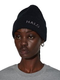 Vrouw met een donkere huid die een zwarte gebreide muts draagt met de tekst "HALO", zilveren oorringen en een grijs T-shirt met ronde hals, kijkt recht vooruit met een neutrale uitdrukking.