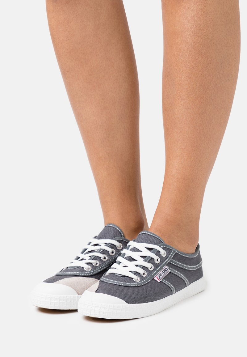 Jambes avec des baskets basses en toile grise, lacets blancs et semelles en caoutchouc blanches sur un fond clair.