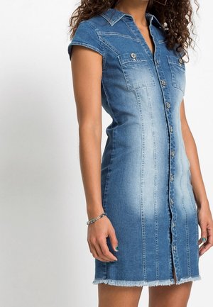 Donna che indossa un abito aderente in denim a maniche corte con bottoni sul davanti, tasche sul petto, orlo sfilacciato e braccialetto d'argento al polso sinistro.