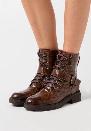 Bottines en cuir marron à motif croco avec lacets, fermetures éclair latérales, sangles à boucle et semelles épaisses en caoutchouc noir portées sur des jambes nues.