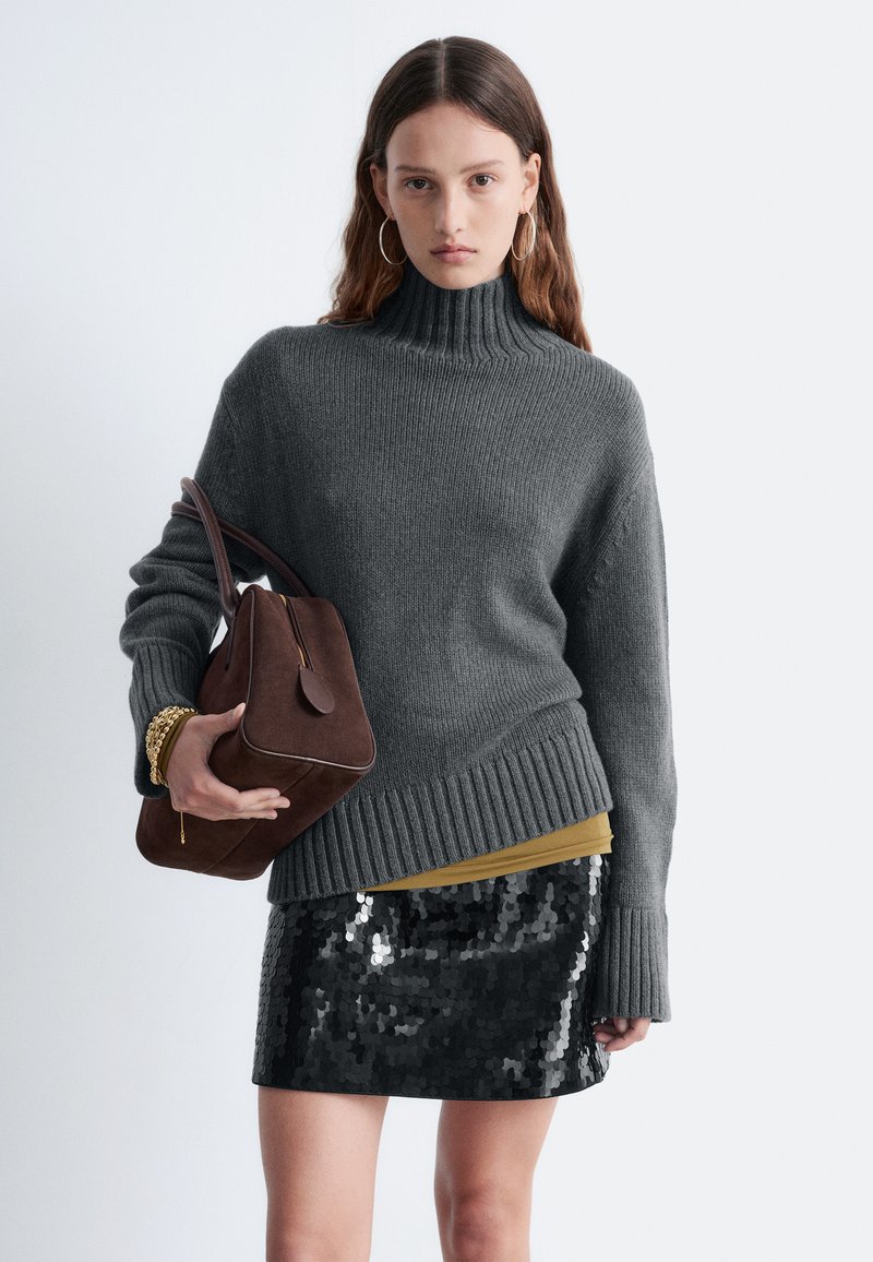 Un pull en tricot gris avec un col roulé haut, superposé sur un haut moutarde, assorti à une mini jupe noire en sequins et un sac à main marron.