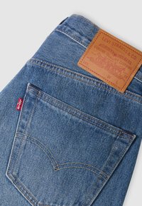 Jean en denim bleu plié montrant une poche arrière avec des coutures délavées, une étiquette en cuir marron de la marque Levi's et une petite étiquette rouge.
