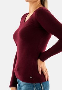 Maglione bordeaux a maniche lunghe con scollatura a V realizzato in tessuto morbido, caratterizzato da un design aderente e da un piccolo dettaglio decorativo sul lato inferiore.