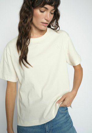 Vrouw met lang golvend haar, gekleed in een effen lichtbeige T-shirt met korte mouwen en een blauwe spijkerbroek, kijkt naar beneden met één hand in haar zak.