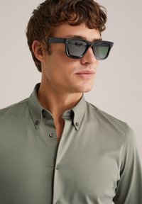 Schwarze quadratische Sonnenbrille mit getönten Gläsern, kombiniert mit einem hellolivgrünen Hemd mit Knöpfen, das einen Kragen und einen dezenten Glanz aufweist.