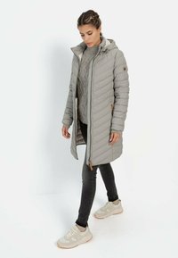 Lichtgrijze, gewatteerde lange jas met een capuchon en een rits aan de voorkant; daaroverheen een grijze gebreide trui. Gepaard met donkere jeans en beige sneakers.