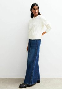 Frau mit dunklen Haaren in einem weißen, grob gestrickten Pullover, weiten blauen Jeans und schwarzen Schuhen steht an einer weißen Wand und schaut zur Seite.