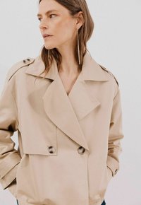 Beige, dubbelknäppt trenchcoat med stora slag, knappdetaljer och lös passform. Mjuk tyg, synlig sömnad, minimal design.