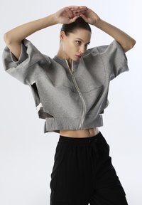 Grijze cropped jas met een rits aan de voorkant, losse mouwen en zijuitsparingen. Gecombineerd met zwarte broek met trekkoord. Soepele stoftextuur.