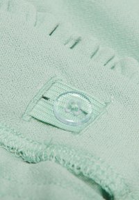 Mintunvihreä kangas, jossa on teksturoidun pinnan lisäksi kirkas muovinappi ja pieni, kiinnitetty etiketti, jonka reunat ovat röyhelöidyt.