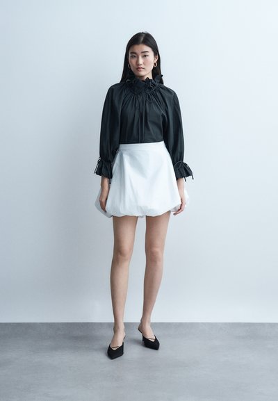 Νεαρή γυναίκα που φοράει μια μαύρη μπλούζα με μακριά μανίκια, βολάν στους μανσέτες και το γιακά, μια λευκή φούστα με όγκο και μαύρες μυτερές γόβες, στέκεται σε εσωτερικό χώρο.