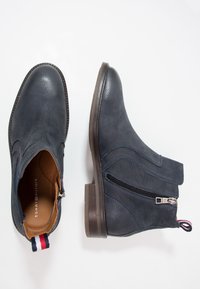 Tmavě modré kožené kotníkové boty mají boční zip, texturovaný povrch a hnědý podpatek. Obsahují červenou, bílou a modrou poutko.