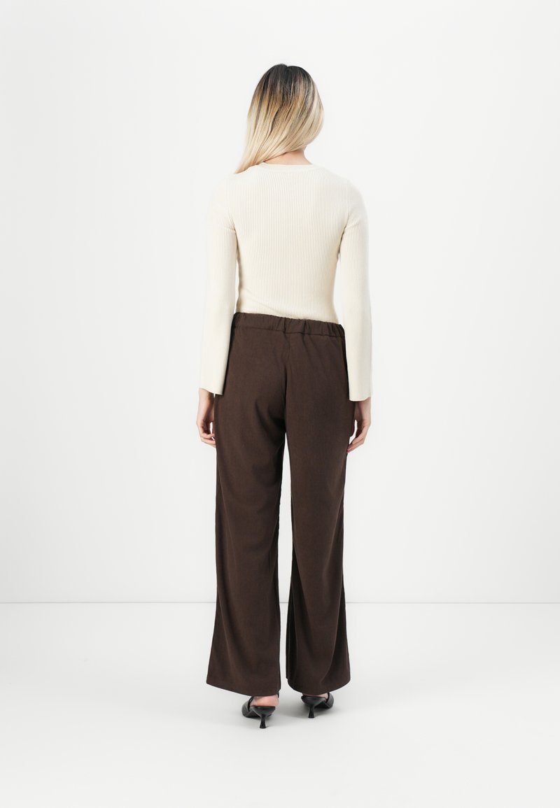 Vrouw met lang blond haar, gekleed in een crèmekleurig, geribbeld, lang mouwtop, een bruin wijde broek en zwarte hoge hakken, met haar gezicht van de camera af, tegen een witte achtergrond.