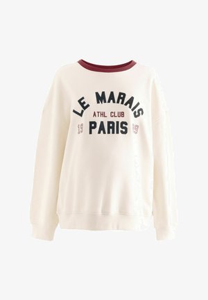 Kremeni dukserica s burgundskim ovratnikom i rebrastim manžetama. Sadrži natpis "LE MARAIS ATHL CLUB PARIS" u crnom i burgundskom slovlju. Opisuje ležeran kroj.