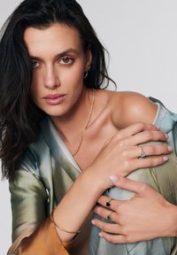 Złoty naszyjnik, wiele pierścionków z niebieskimi, zielonymi i czarnymi kamieniami szlachetnymi. Modelka nosi zwiewną, wielokolorową satynową bluzkę o luźnym kroju.