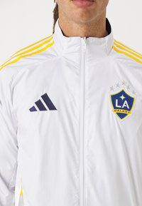 Chaqueta blanca con cremallera y acentos en amarillo y marino, que presenta el logo de Adidas en el lado izquierdo del pecho y el emblema de LA Galaxy con estrellas. Textura suave.